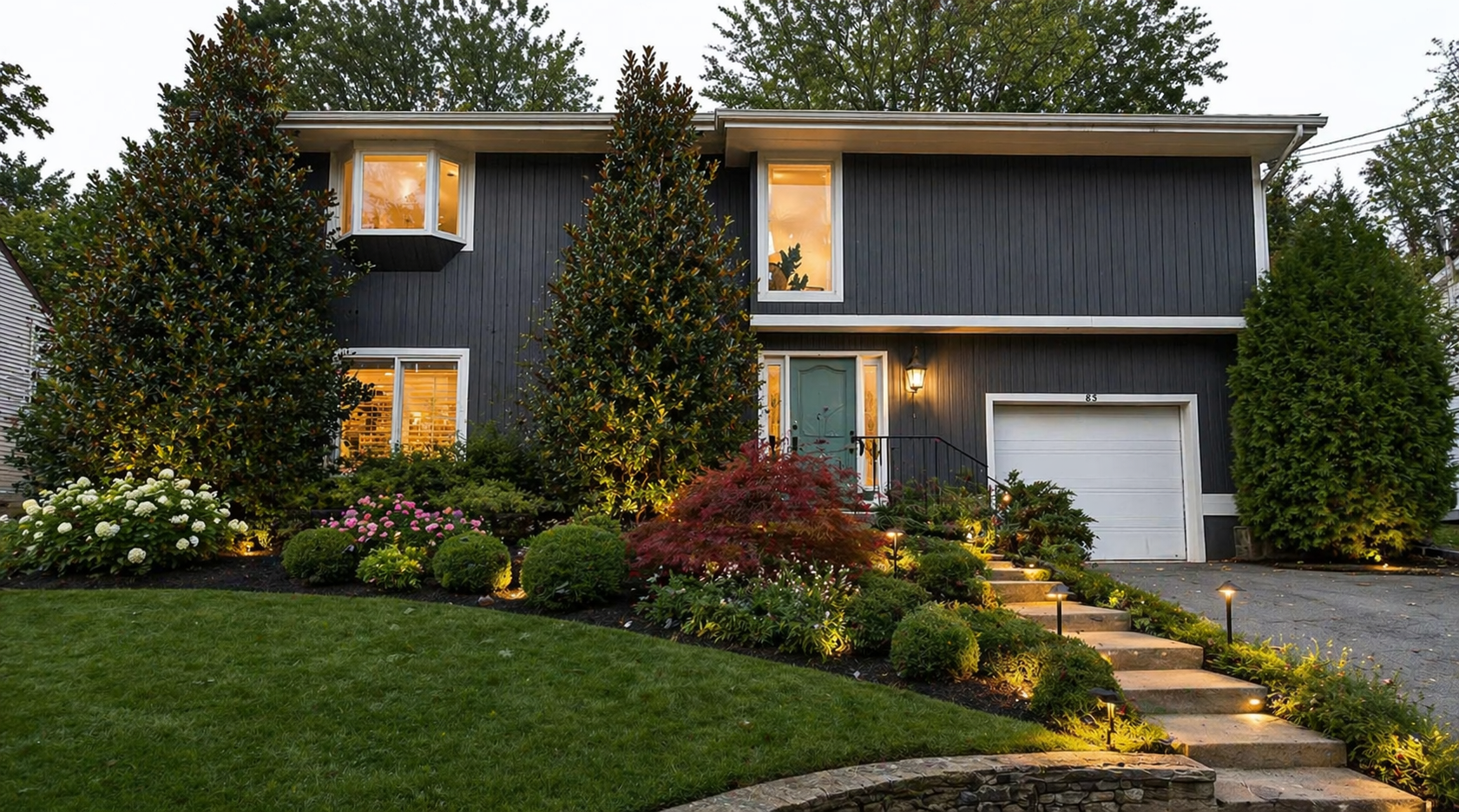 Staged home exterior at twilight with landscape lighting and lush gardens
